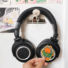 Black over-ear headphones with a cartoon character sticker on the right earcup, hanging on a white wall with posters in the background and a hand adjusting the sticker.