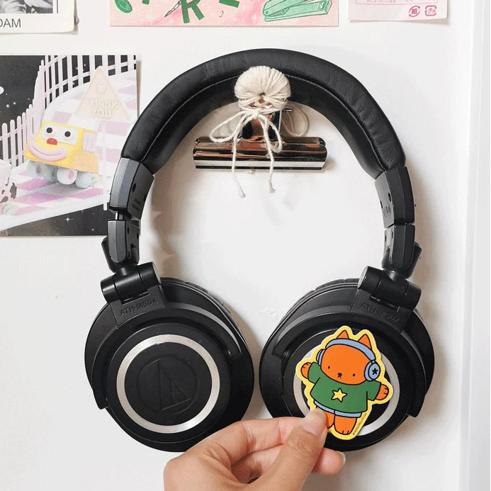 Black over-ear headphones with a cartoon character sticker on the right earcup, hanging on a white wall with posters in the background and a hand adjusting the sticker.