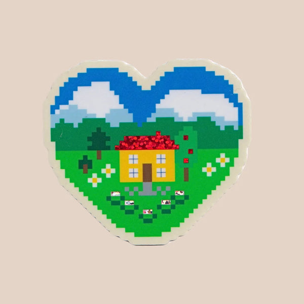 A heart-shaped sticker featuring a pixel-art landscape with a yellow house, red roof, green fields, trees, flowers, and blue sky, shown on a plain beige background.