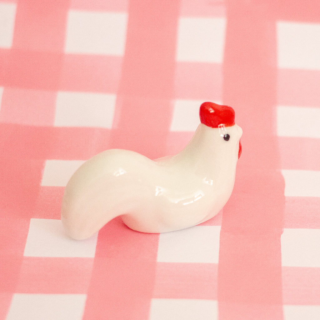 A glossy white ceramic chicken figurine with a bright red comb, standing on a pink and white checkered surface.