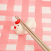 A small glossy white chicken-shaped chopstick holder with red comb and wattle detail, holding two metallic chopsticks, set against a pink and white checkered background.