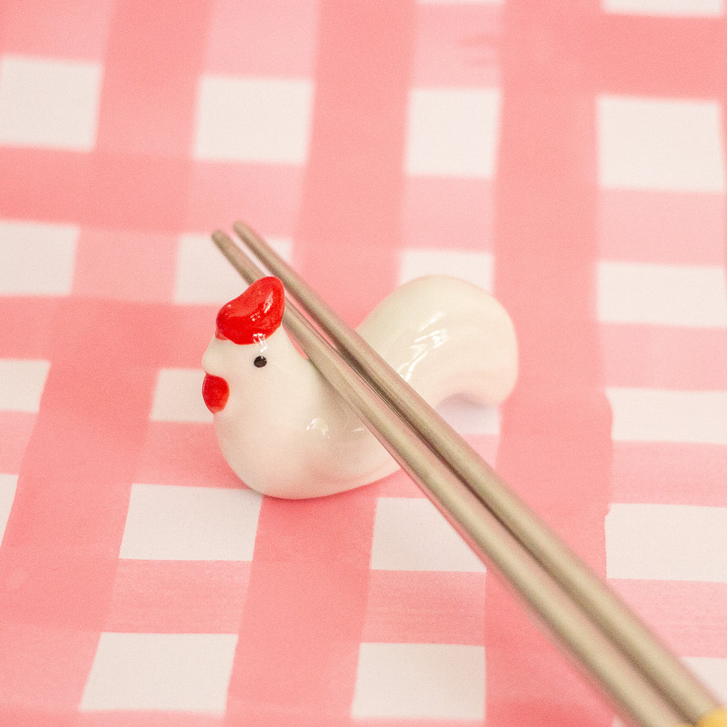 A small glossy white chicken-shaped chopstick holder with red comb and wattle detail, holding two metallic chopsticks, set against a pink and white checkered background.