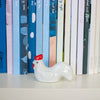 A glossy white ceramic chicken figurine with a red comb and wattle, placed on a white shelf in front of a row of colourful books.