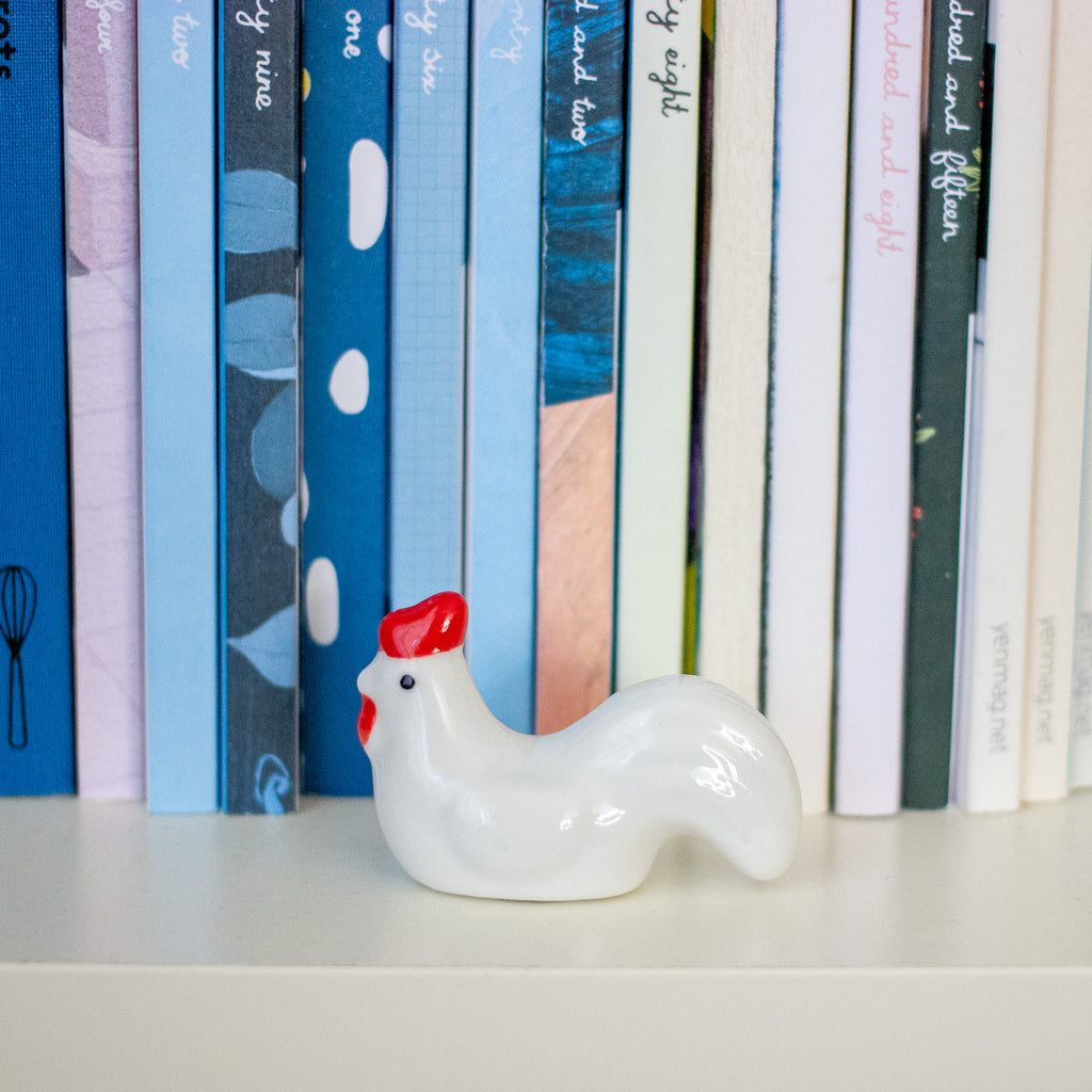 A glossy white ceramic chicken figurine with a red comb and wattle, placed on a white shelf in front of a row of colourful books.