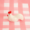 A small glossy white chicken figurine with a bright red comb and red beak, standing on a pink-and-white checkered surface.