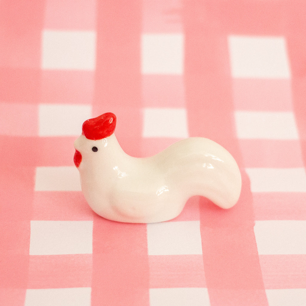 A small glossy white chicken figurine with a bright red comb and red beak, standing on a pink-and-white checkered surface.