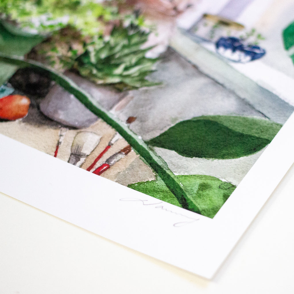 A close-up of a watercolour art print featuring green leaves, succulents, and a stone garden scene, with a wide white border and a signature at the bottom.