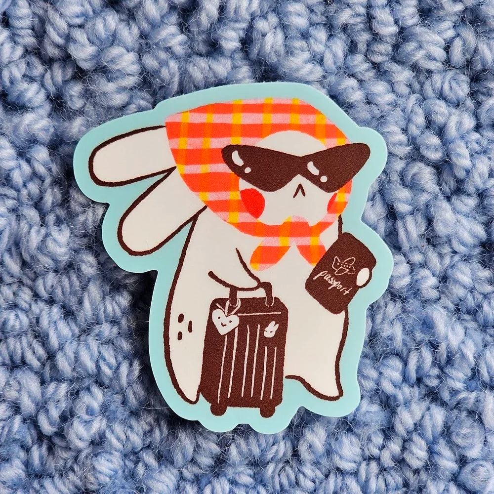 A die-cut sticker featuring a cute cartoon bunny character wearing sunglasses, a red and orange plaid scarf, and holding a brown suitcase, placed on a textured blue-grey background.