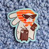 A die-cut sticker featuring a cute cartoon bunny character wearing sunglasses, a red and orange plaid scarf, and holding a brown suitcase, placed on a textured blue-grey background.
