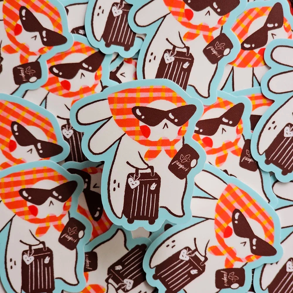 A pile of die-cut stickers featuring a cartoon rabbit wearing black sunglasses, an orange and white plaid scarf, and holding a suitcase, with a blue outline. 