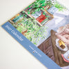 A spiral-bound 2027 desk calendar titled 'Home and Cats' by Nanan Studio, featuring a watercolour illustration of a ginger cat sitting on a wooden table next to a lamp and plants.