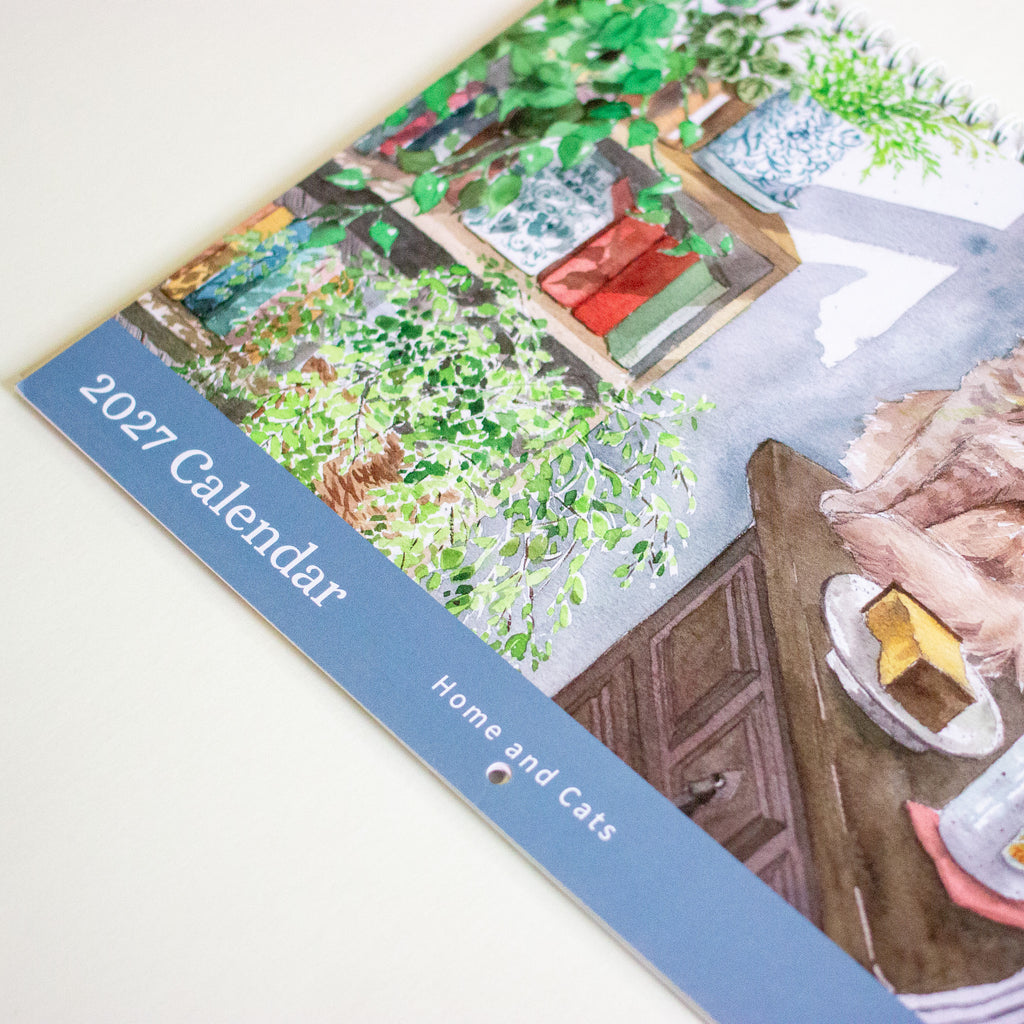 A spiral-bound 2027 desk calendar titled 'Home and Cats' by Nanan Studio, featuring a watercolour illustration of a ginger cat sitting on a wooden table next to a lamp and plants.