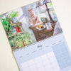 A spiral-bound wall calendar page shown at an angle on a light beige background. The top portion features a watercolour illustration of a ginger cat sitting on a wooden table in a cosy room with plants, a lamp, and a mug.