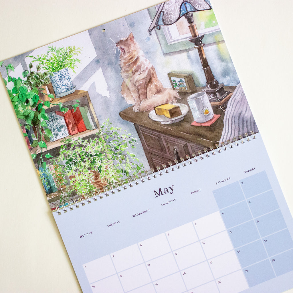 A spiral-bound wall calendar page shown at an angle on a light beige background. The top portion features a watercolour illustration of a ginger cat sitting on a wooden table in a cosy room with plants, a lamp, and a mug.