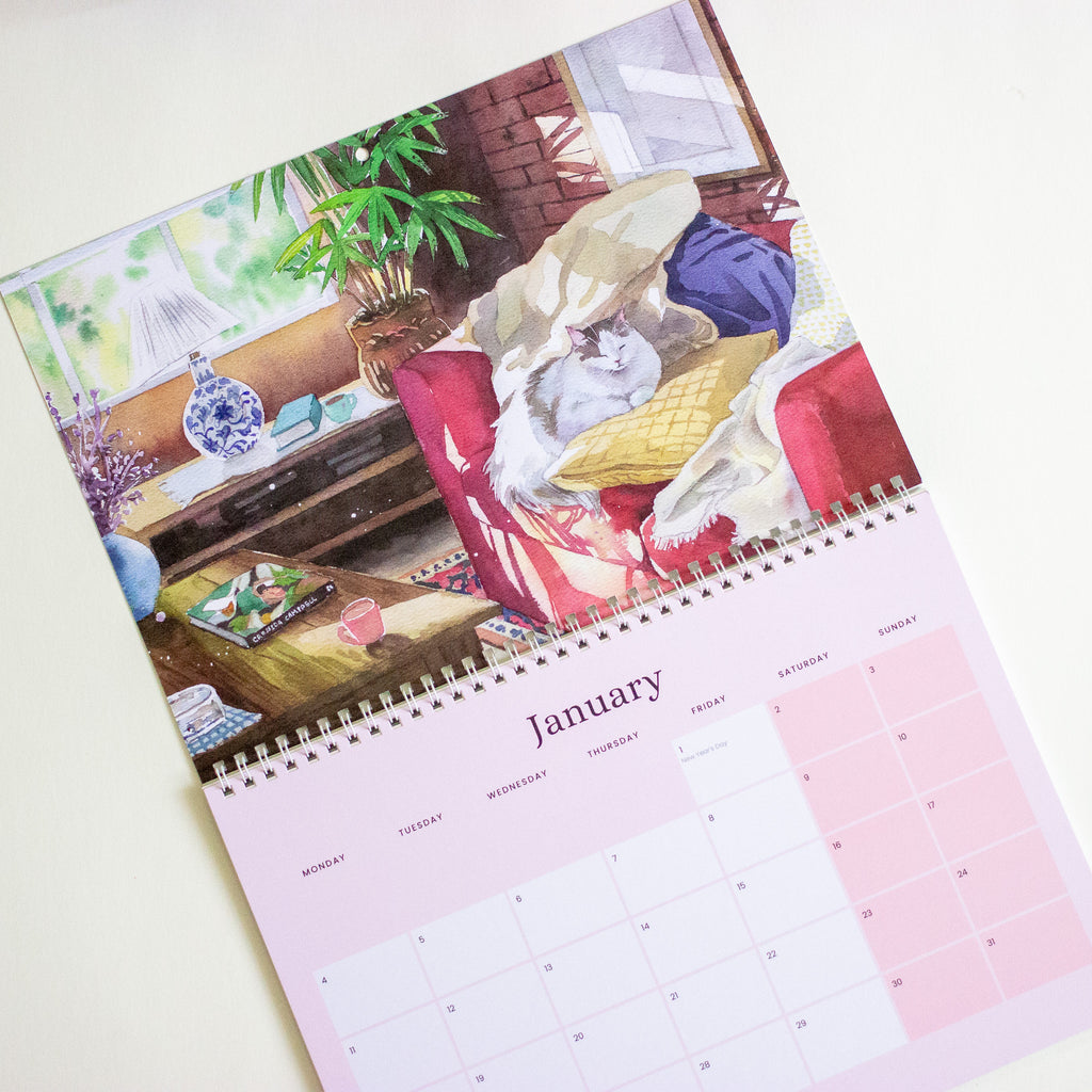 A spiral-bound wall calendar page shown at an angle on a light beige background. The top portion features a watercolour illustration of a cat sitting on a sofa in a cosy room with plants, a lamp, and a mug.