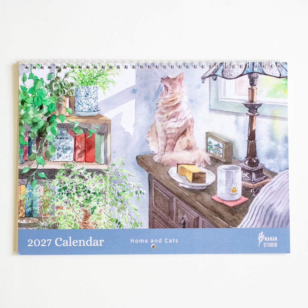 A spiral-bound 2027 desk calendar titled 'Home and Cats' by Nanan Studio, featuring a watercolour illustration of a ginger cat sitting on a wooden table next to a lamp and plants.