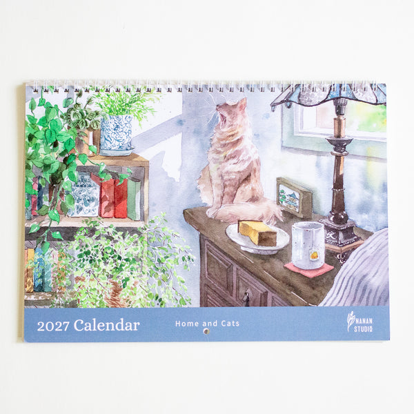 A spiral-bound 2027 desk calendar titled 'Home and Cats' by Nanan Studio, featuring a watercolour illustration of a ginger cat sitting on a wooden table next to a lamp and plants.