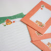 A close-up of stationery featuring lined writing paper and matching red-orange envelopes with a cute teddy bear illustration and a green snow-themed strip.