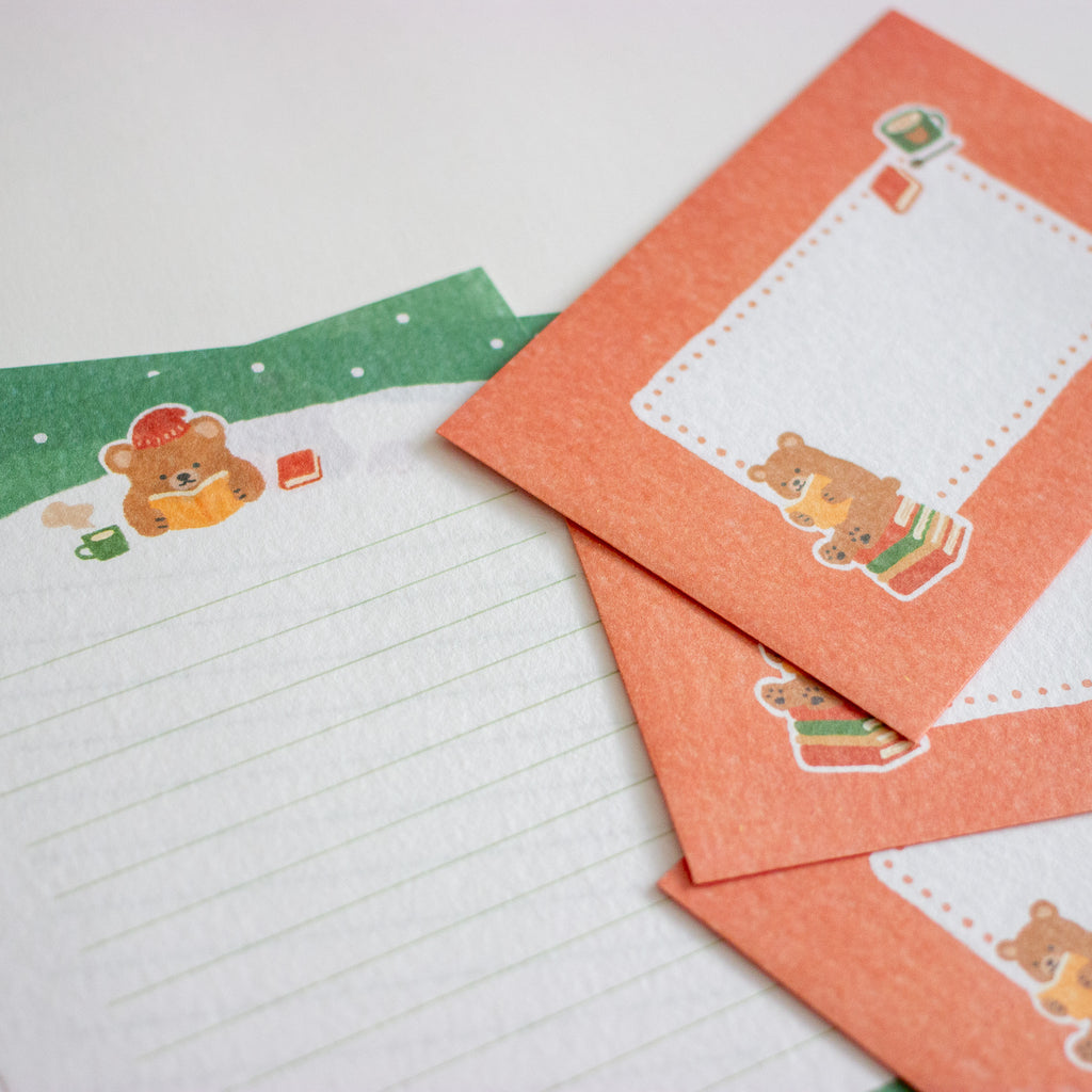 A close-up of stationery featuring lined writing paper and matching red-orange envelopes with a cute teddy bear illustration and a green snow-themed strip.
