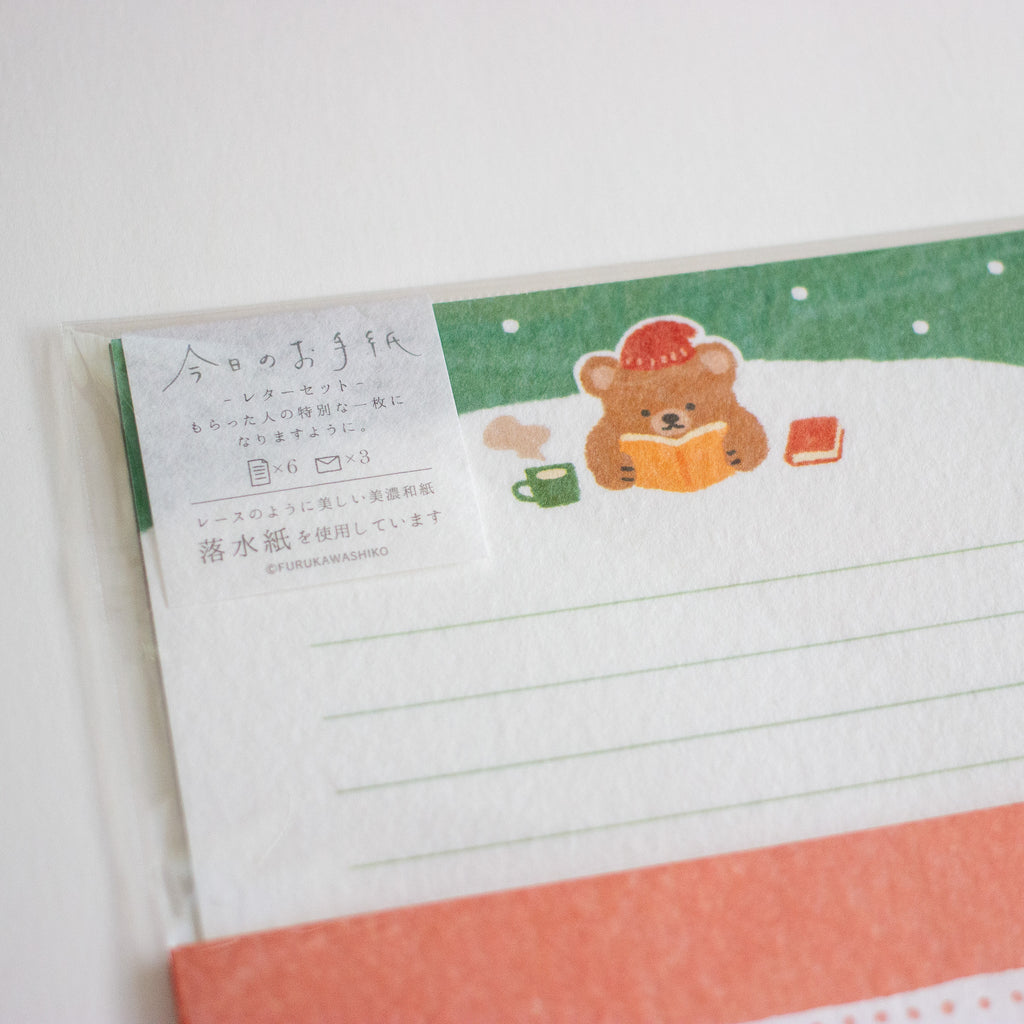 A close-up of a Japanese stationery card or letterhead with a cute teddy bear illustration reading a book in the snow, green header band, lined writing area, and a coral-orange border at the bottom.