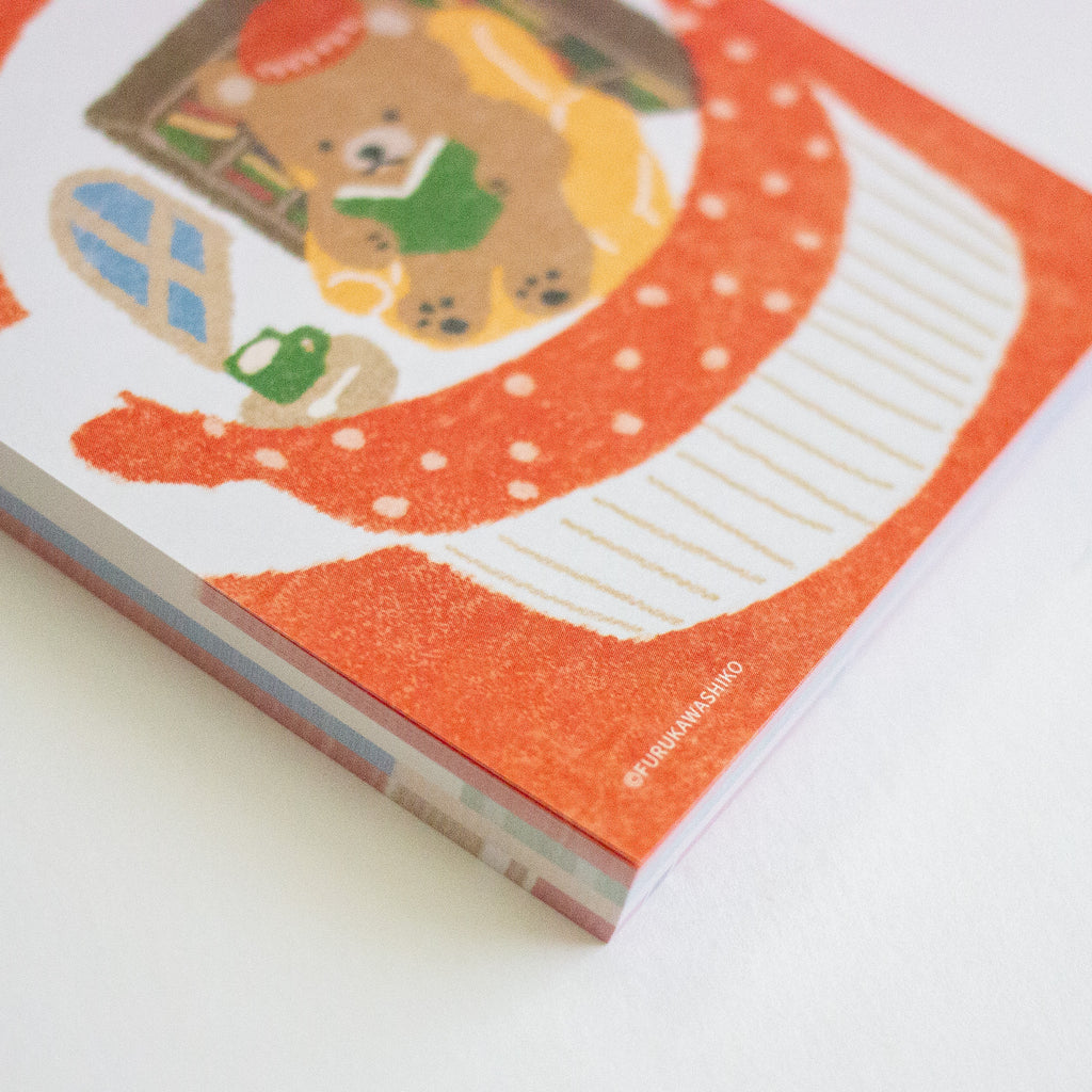 A close-up of a square printed memo pad cover featuring a teddy bear illustration and a large orange-and-white curved graphic, photographed on a white background.