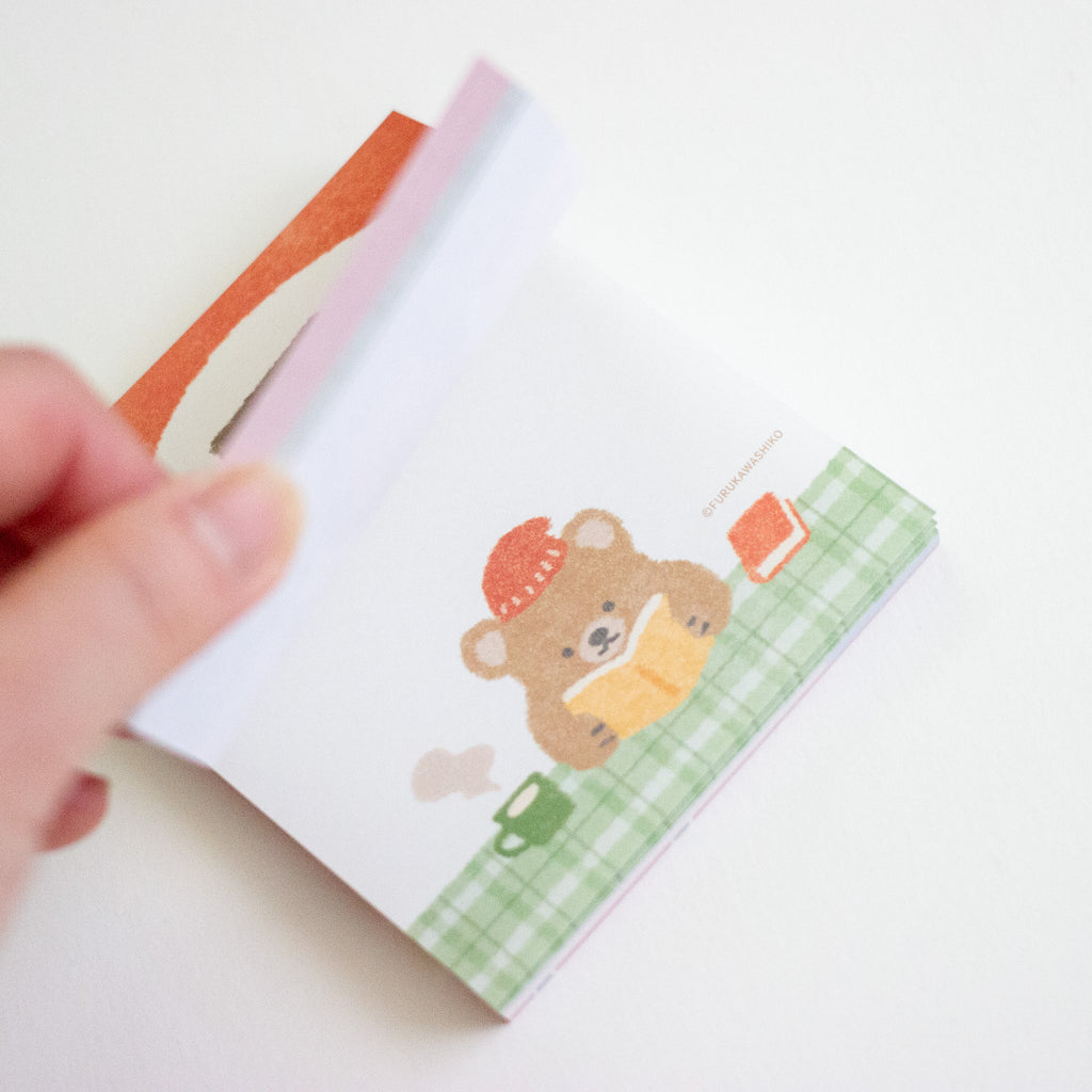 A hand holding a small memo pad featuring a cute cartoon bear reading a book, with a green plaid pattern and orange accents, on a plain white background.