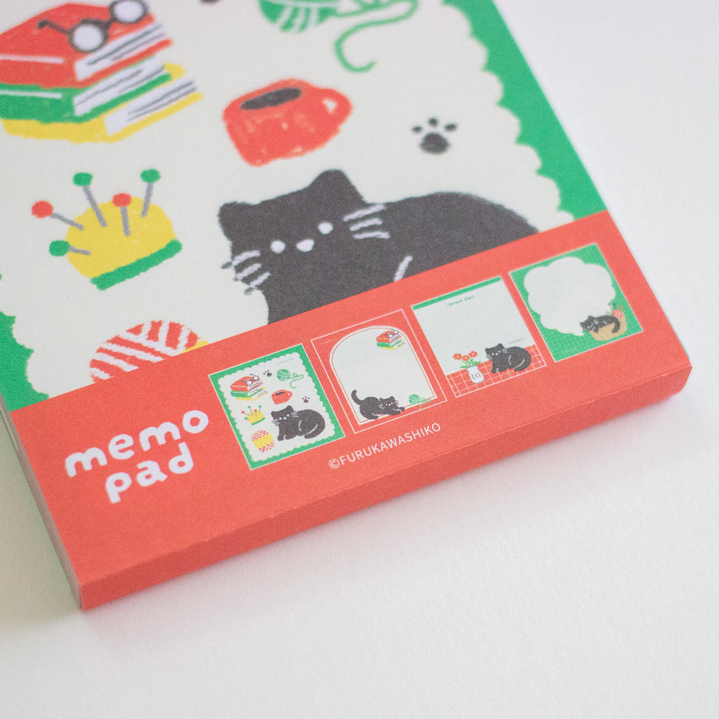 A close-up of a memo pad package featuring a white background with a black cat illustration, colourful books and cup graphics, and a coral-red lower band labeled 'memo pad'.