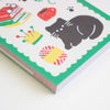 A close-up of a colourful memo pad with a white background, green border, and playful illustrations including a black cat, red and yellow objects, and small paw prints.