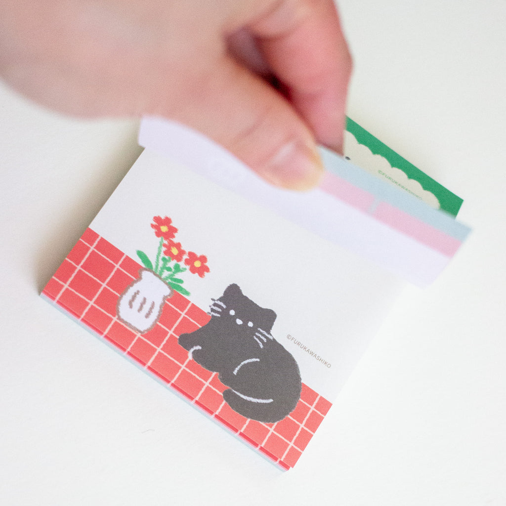 A hand is holding a small printed memo pad featuring a black cat sitting on a red grid pattern next to red flowers in a white pot. 