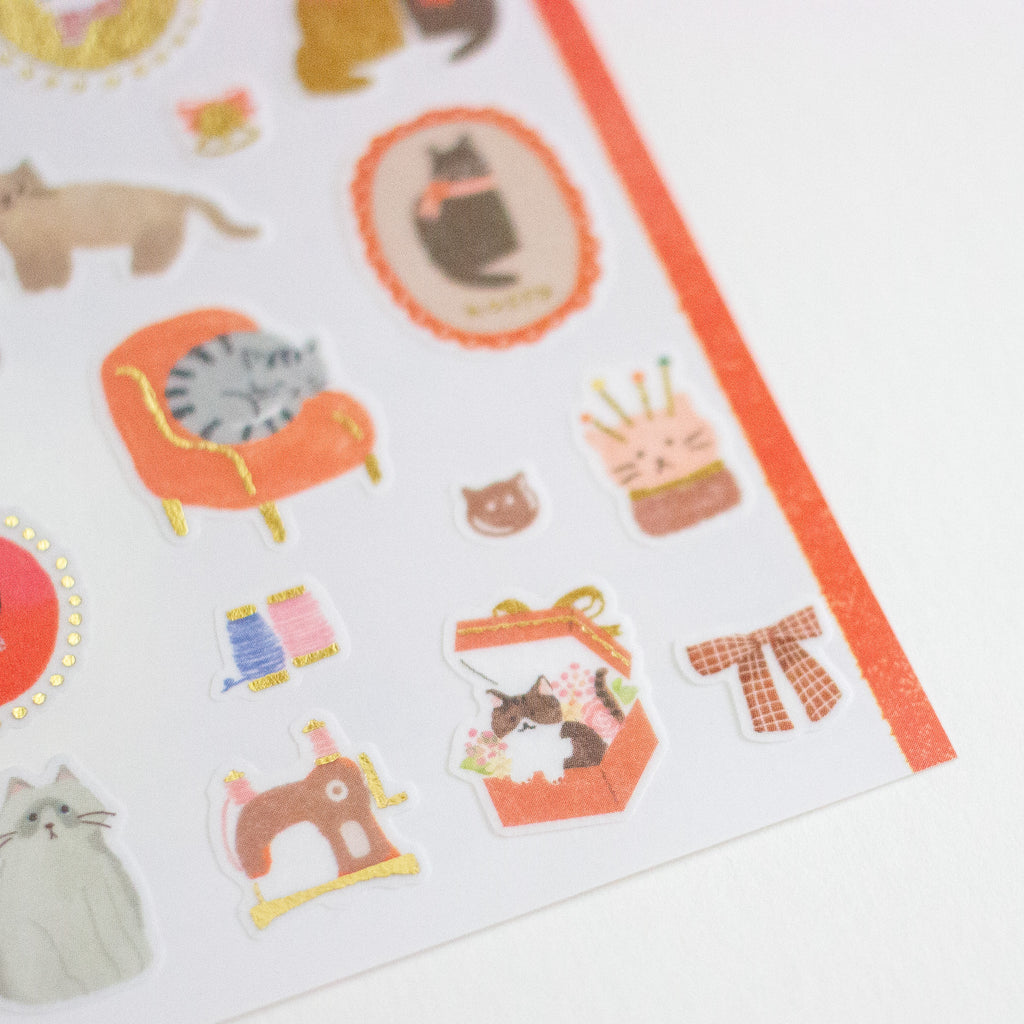 A close-up of a sheet of cute cat-themed stickers on a white background, featuring illustrations of cats, an orange armchair, a sewing machine, and small accessories.
