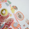A close-up of a hand holding a sheet of cute cat-themed stickers, including gold foil circular designs, pastel cat illustrations, and small decorative motifs on a white background.