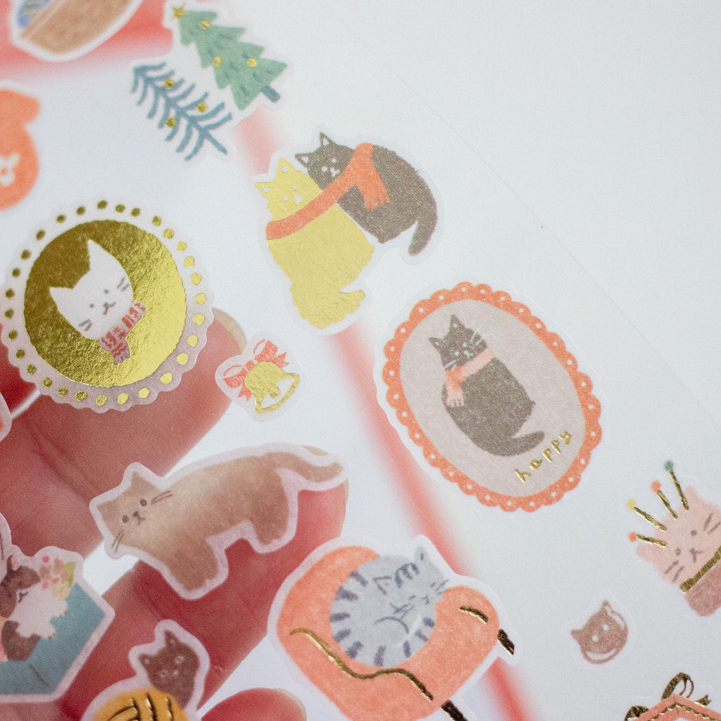 A close-up of a hand holding a sheet of cute cat-themed stickers, including gold foil circular designs, pastel cat illustrations, and small decorative motifs on a white background.