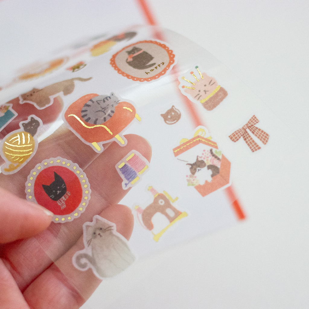 A hand holding a clear roll of decorative stickers featuring cute cat-themed illustrations, including a cat in a photo frame, cat furniture, and cat accessories, against a clean white background.