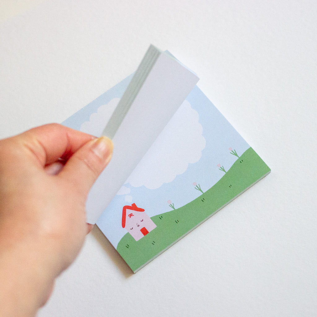 A hand is holding a stack of square notepad sheets featuring a cute illustration of a small house on a green hill under a blue sky.