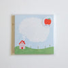 A square memo pad in clear plastic wrapping, featuring a light blue sky, green hill, small pink house, and a large blank white speech bubble. A red apple sticker is in the top right corner.