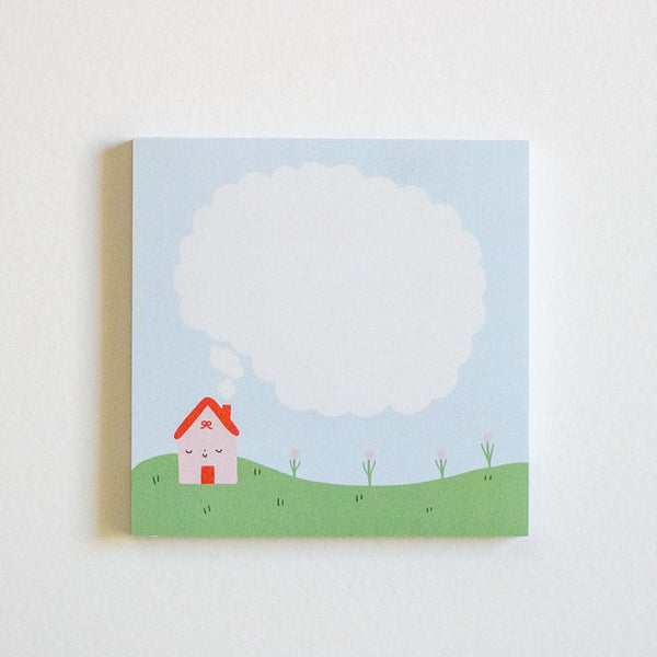 A square notepad with a whimsical illustration of a small house on a green hill under a large blank speech bubble, set against a plain white background.