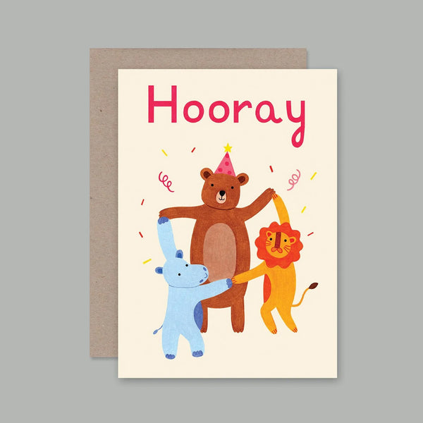 A greeting card with a kraft envelope, featuring the word 'Hooray' in pink above an illustration of a brown bear, a yellow lion, and a blue hippo dancing together on a light grey background.