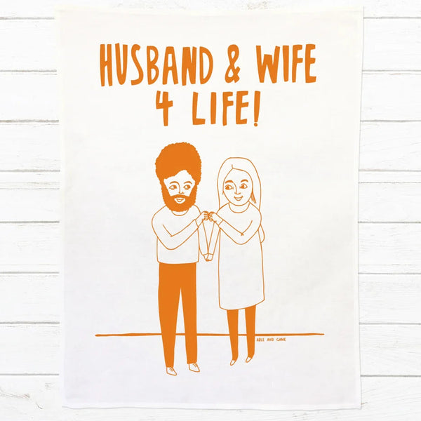 A white fabric tea towel or hanging with orange printed text reading 'HUSBAND & WIFE 4 LIFE!' and a simple orange line illustration of a bearded man and a woman holding hands, displayed against a white wood plank background.