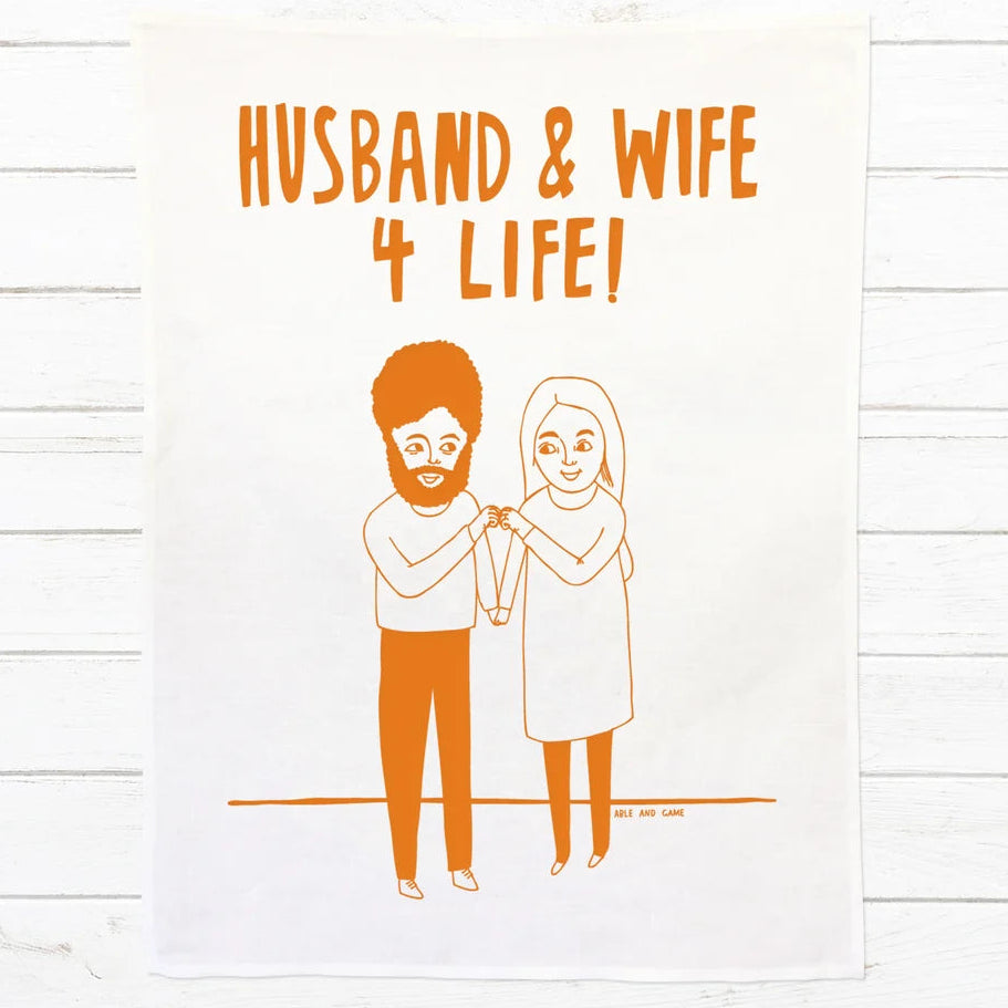 A white fabric tea towel or hanging with orange printed text reading 'HUSBAND & WIFE 4 LIFE!' and a simple orange line illustration of a bearded man and a woman holding hands, displayed against a white wood plank background.