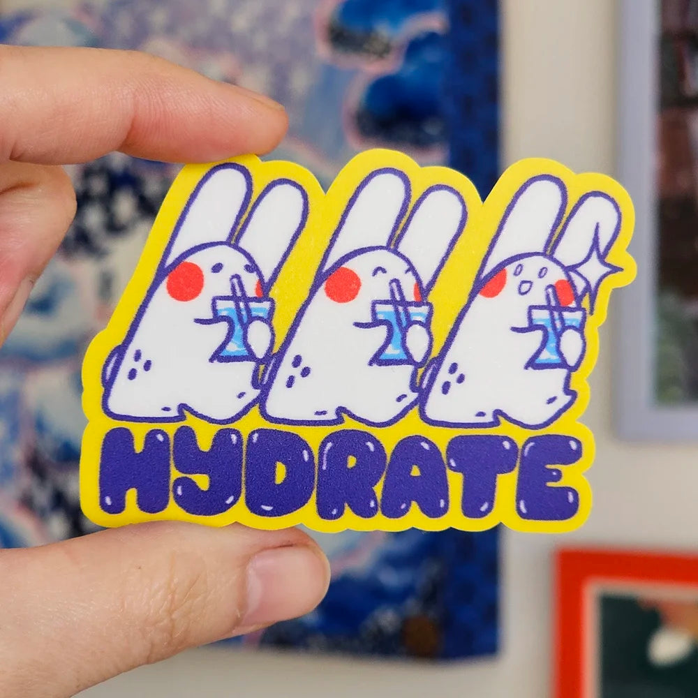 A hand holds a die-cut sticker featuring three cartoon white rabbits with blue drink glasses and red cheeks, outlined in yellow, with the word 'HYDRATE' in blue bubbly letters at the bottom. The background is a blurred indoor setting with framed art.