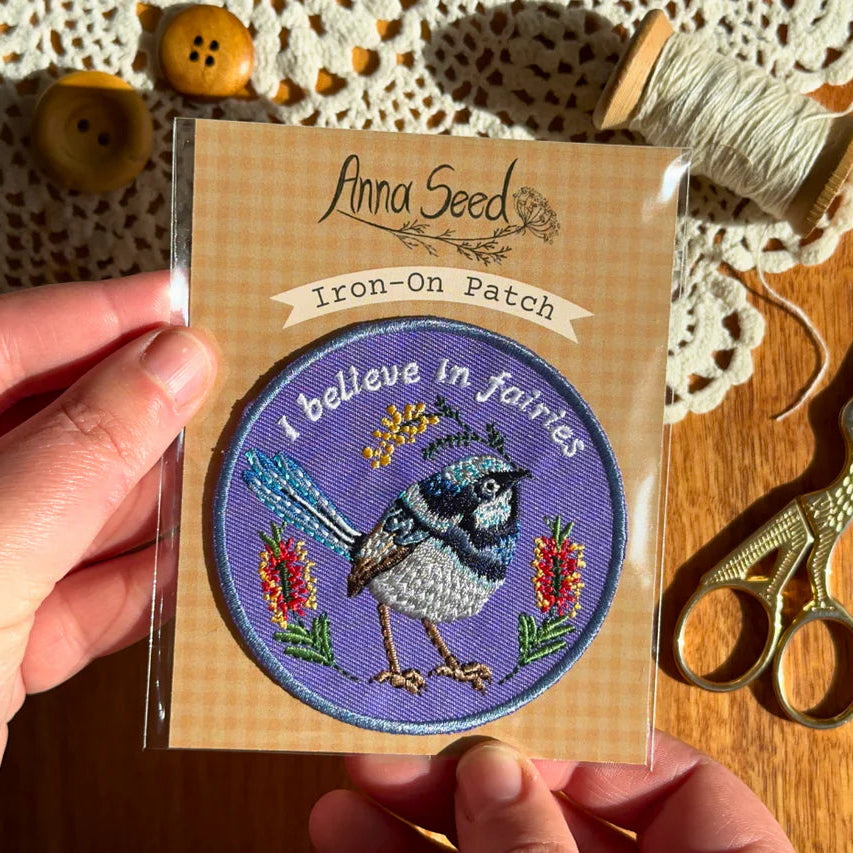 Iron-on patch with bird design and 'I believe in fairies' text, held by a hand on a wooden surface with sewing items.