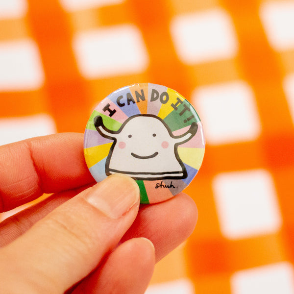 A close-up of a hand holding a round pinback button badge featuring a cartoon character and the text 'I CAN DO IT!!' against a blurred orange and white checkered background.