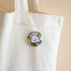 A round pin-back button featuring a white cartoon with the words 'I CAN DO IT' on a colourful background, pinned onto a white canvas tote bag against a soft beige background.