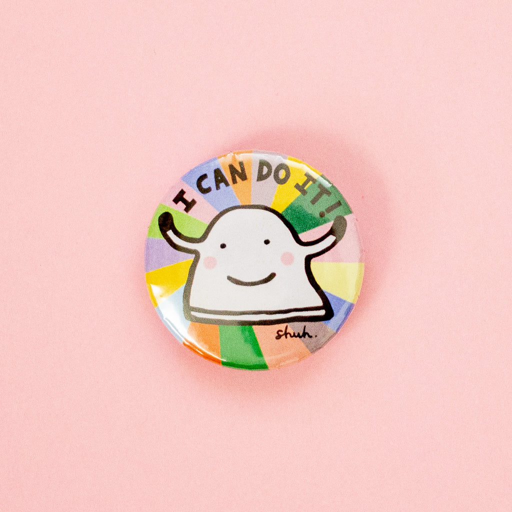 A round pinback button badge featuring a cartoon sheep character with the text 'I CAN DO IT!' and a small 'shuh.' signature, set against a colourful rainbow-striped background on a soft pink backdrop.