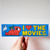 A hand holds a rectangular blue sticker that reads 'I [heart] THE MOVIES.' in yellow letters, with a red cartoon character and a white animal illustration on the left, against a plain light background.