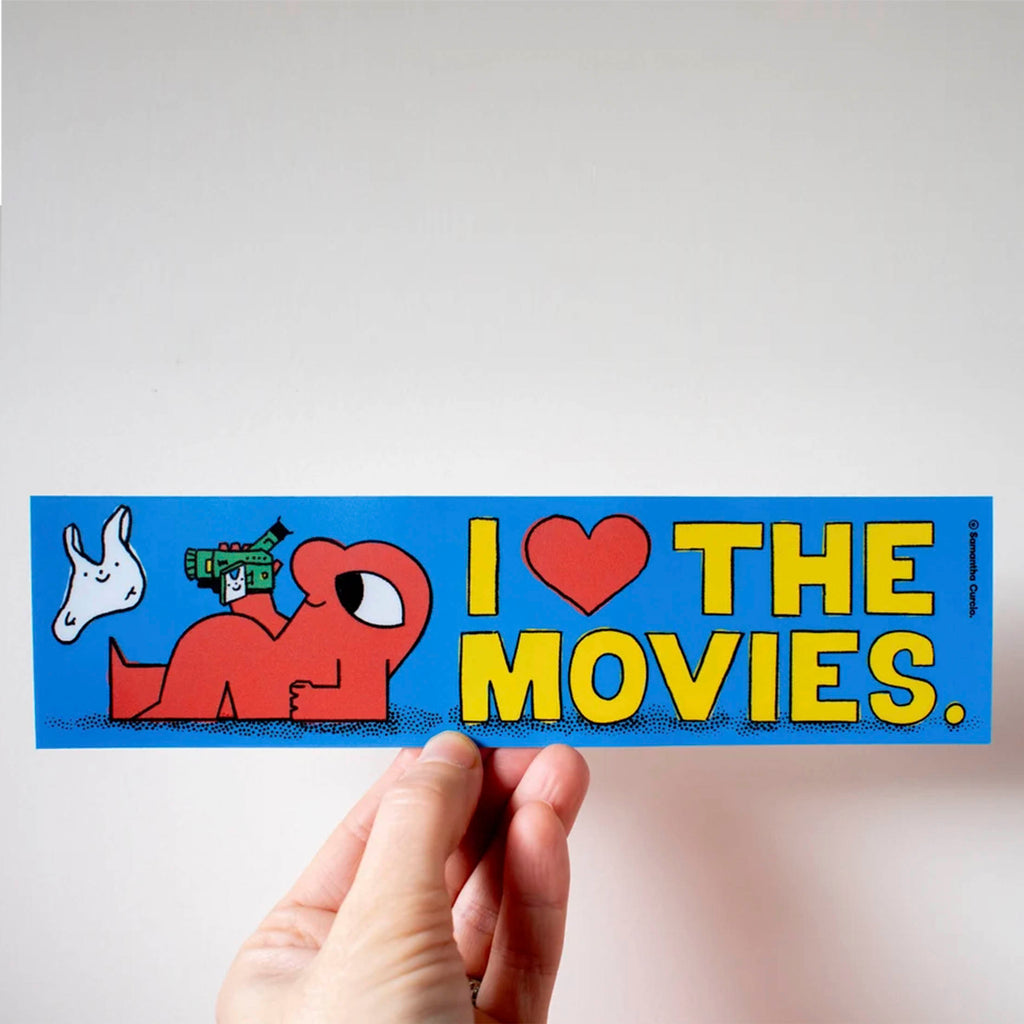A hand holds a rectangular blue sticker that reads 'I [heart] THE MOVIES.' in yellow letters, with a red cartoon character and a white animal illustration on the left, against a plain light background.