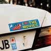 A rectangular bumper sticker with the text 'I [heart] THE MOVIES.' in yellow on a blue background, applied to the rear bumper of a white car.