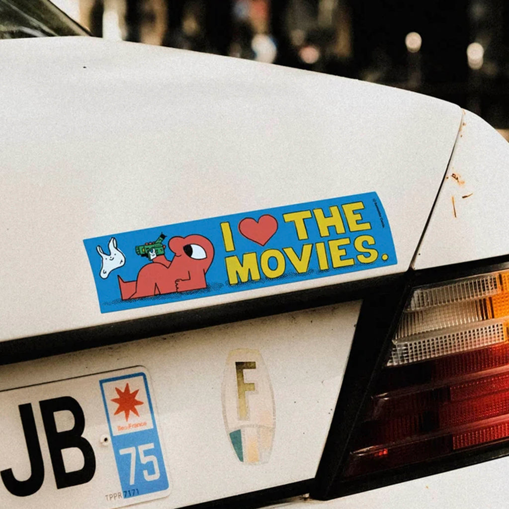 A rectangular bumper sticker with the text 'I [heart] THE MOVIES.' in yellow on a blue background, applied to the rear bumper of a white car.