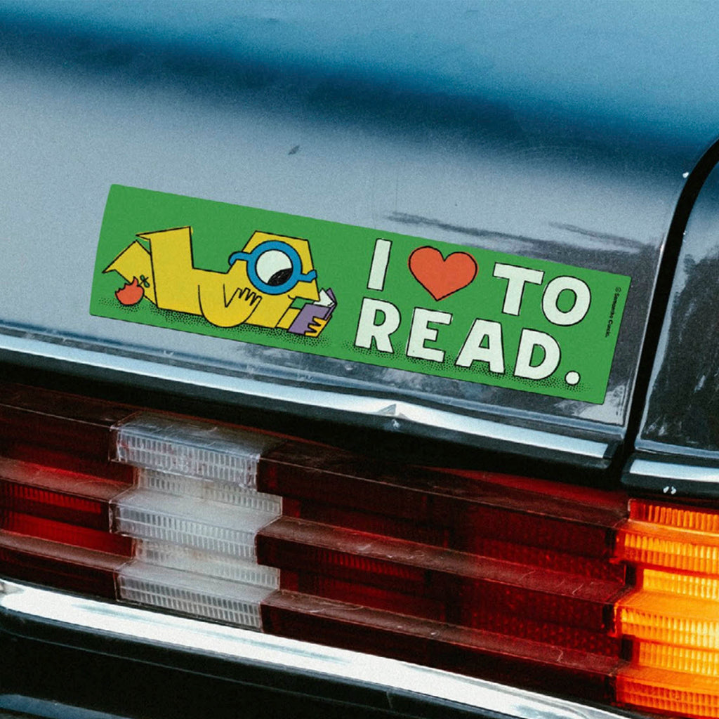 A rectangular bumper sticker with the phrase 'I [heart] TO READ.' in white text on a green background, featuring a cartoon yellow character reading, applied to the rear bumper of a dark car.