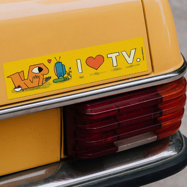 A rectangular yellow car sticker with the text 'I ♥ TV.' and cartoon-style lettering, applied to the rear of a yellow vehicle above the red tail light.